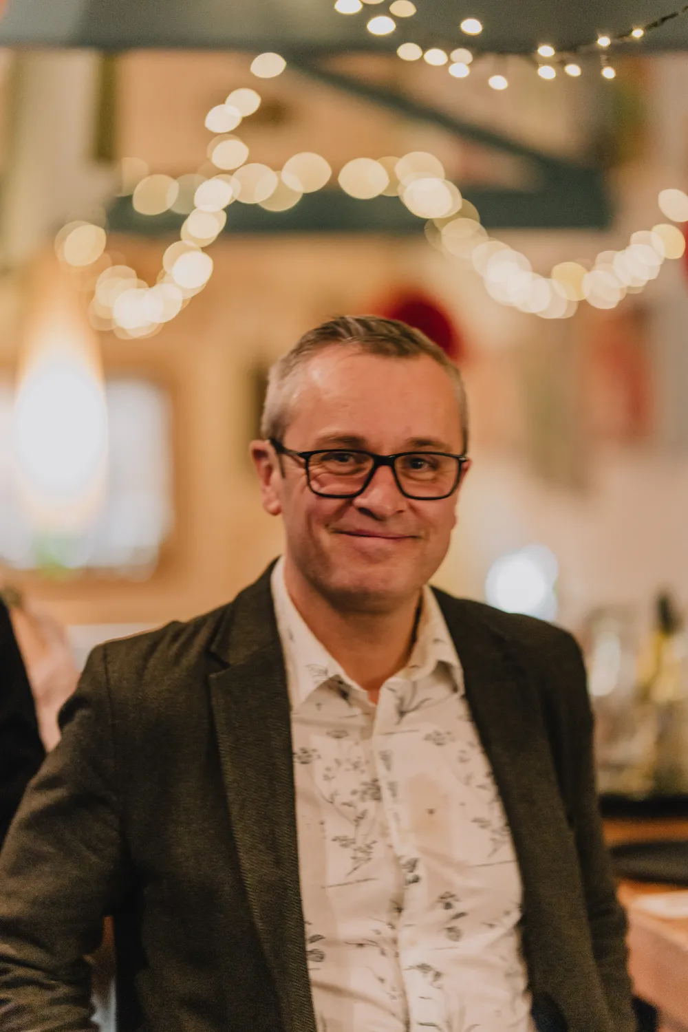 Malcolm smiling at the camera in a nice shirt and jacket with lights in the background